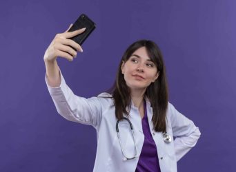 pleased young female doctor in medical robe with stethoscope holds phone up and looks at phone on isolated violet background